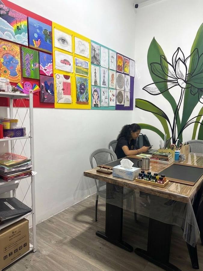 Art room wall covered in student paintings and drawings, with a student at the worktable